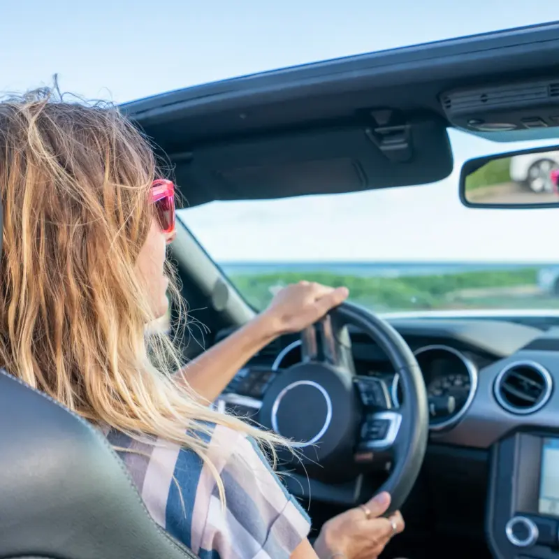 A woman wearing red sunglasses drives a convertible with the top down on a sunny day, one hand on the steering wheel.