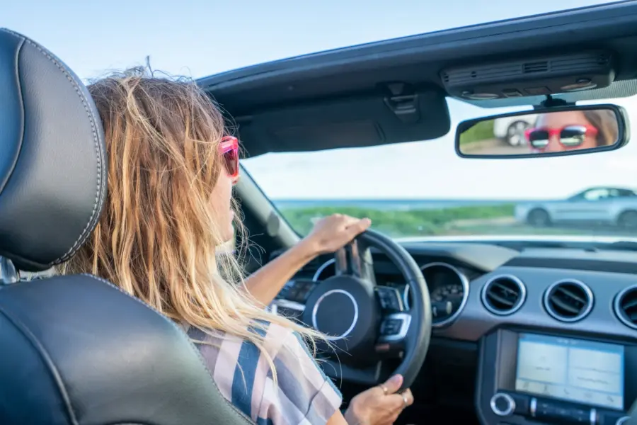 A woman wearing red sunglasses drives a convertible with the top down on a sunny day, one hand on the steering wheel.